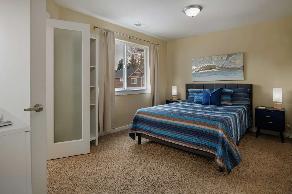 A cozy bedroom with striped bedding, nightstands topped with lamps, and tasteful artwork above the bed. Curtains frame the window, enhancing the sense of privacy and tranquility for restful evenings or leisurely mornings.