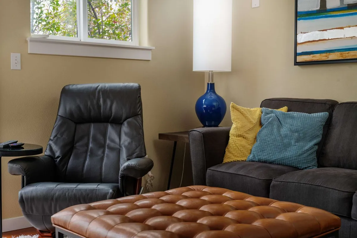 A cozy corner featuring a leather armchair, blue table lamp, colorful cushions, and a tufted coffee table. The ambiance encourages relaxation, reading, or enjoying a cup of coffee, and is well-suited for pet companionship and work breaks.
