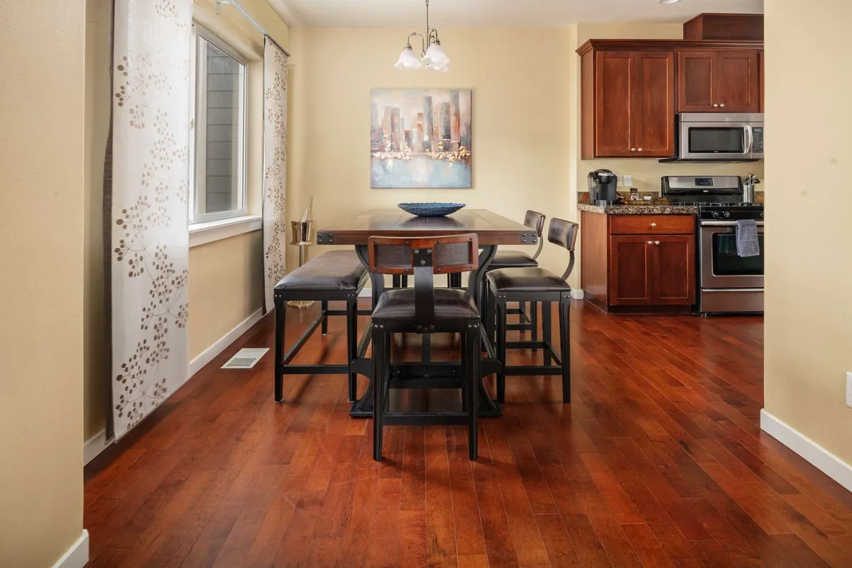 This image features a dining table set in a warm, inviting space with artwork on the wall and a view of the kitchen area. The room is styled for entertaining guests or sharing casual meals, and the atmosphere is cozy and welcoming.