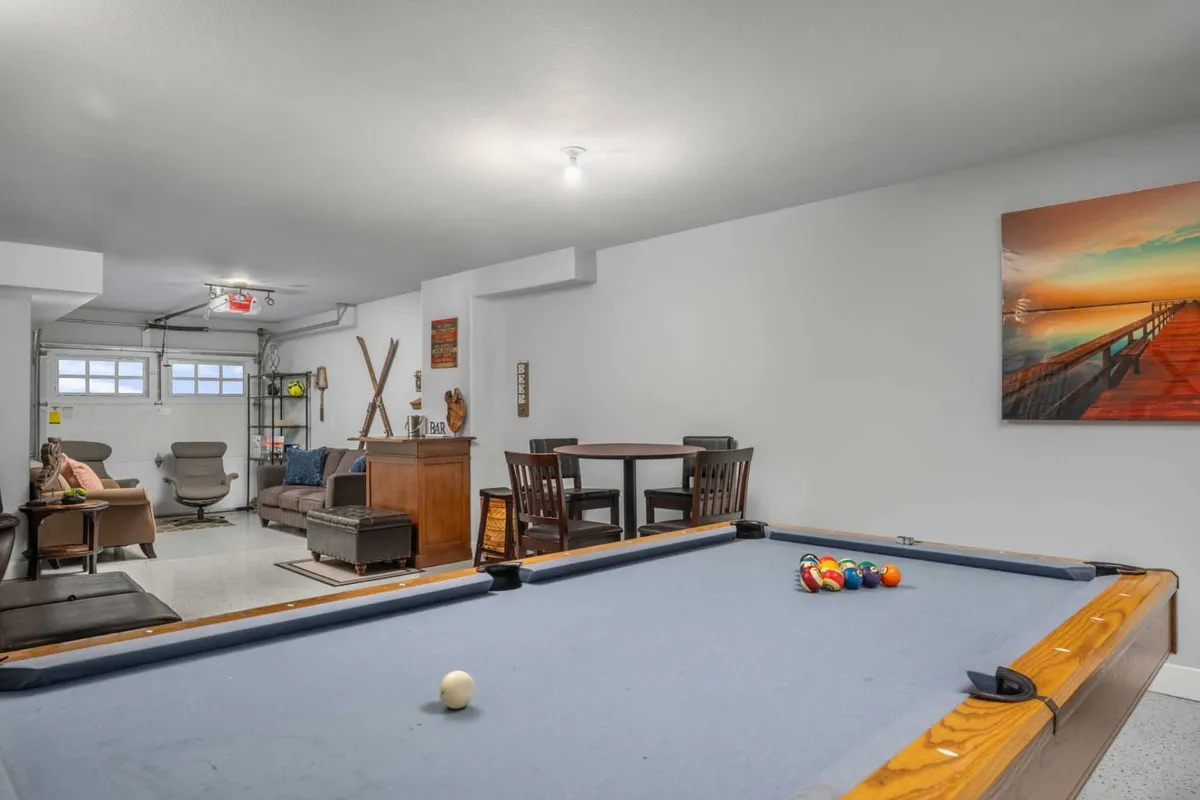 This image captures a lively game room scene, featuring a pool table as the main focal point, a dining table suitable for group gatherings, playful artwork on the wall, and a comfortable seating area. The bright lighting and open layout create an inviting atmosphere for both play and socialization.