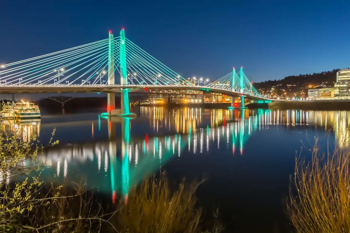 A serene evening scene featuring a bridge over water with city lights reflected on the surface, evoking the peaceful ambiance of Portland nights. The setting suggests opportunities for scenic strolls and family outings in the area.
