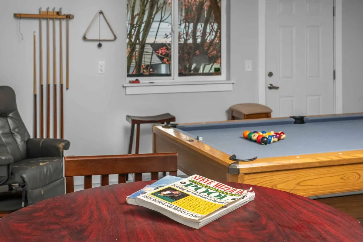 A relaxed recreation lounge featuring a pool table at the center, a wooden table nearby, and a leather chair. The space encourages social interaction with elements for playing pool, reading magazines, or casual relaxation.