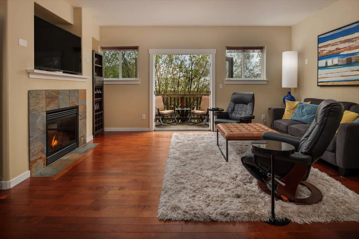 The image showcases a stylish living room with a glowing fireplace as its centerpiece, surrounded by plush seating. Sunlight streams in through open doors that lead to a private balcony, while tasteful artwork adds character to the space.