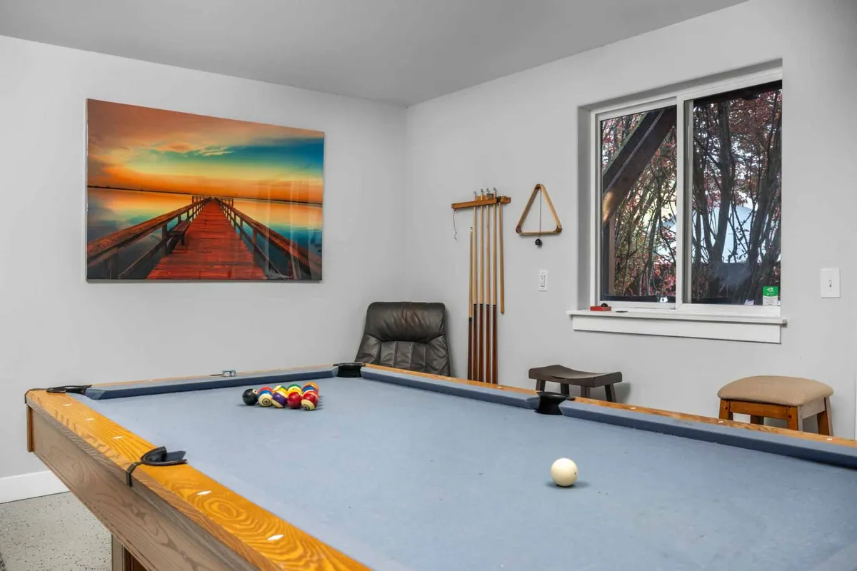 A pool table takes center stage in a cheerful game room, with landscape artwork on the wall and a window offering natural light and glimpses of the outdoors. The setting encourages friendly competition and relaxed conversation.