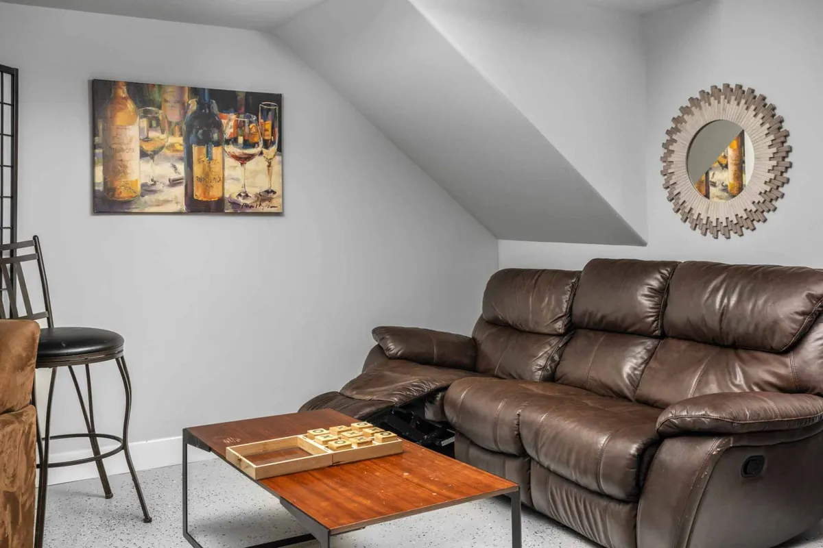 A living area with a brown leather sofa and a wooden coffee table, accented by a framed painting of wine bottles and a decorative mirror. The atmosphere is welcoming and ideal for relaxing or informal gatherings.